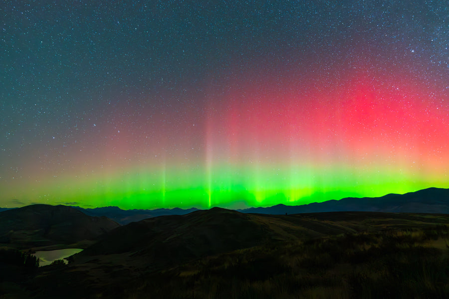 Moccasin Lake Aurora - Metal Print
Rectangular landscape image with a  star-strewn blue sky and bright green and red northern lights appearing over silhouetted hills.  A lake in the lower-left foreground reflects green light.