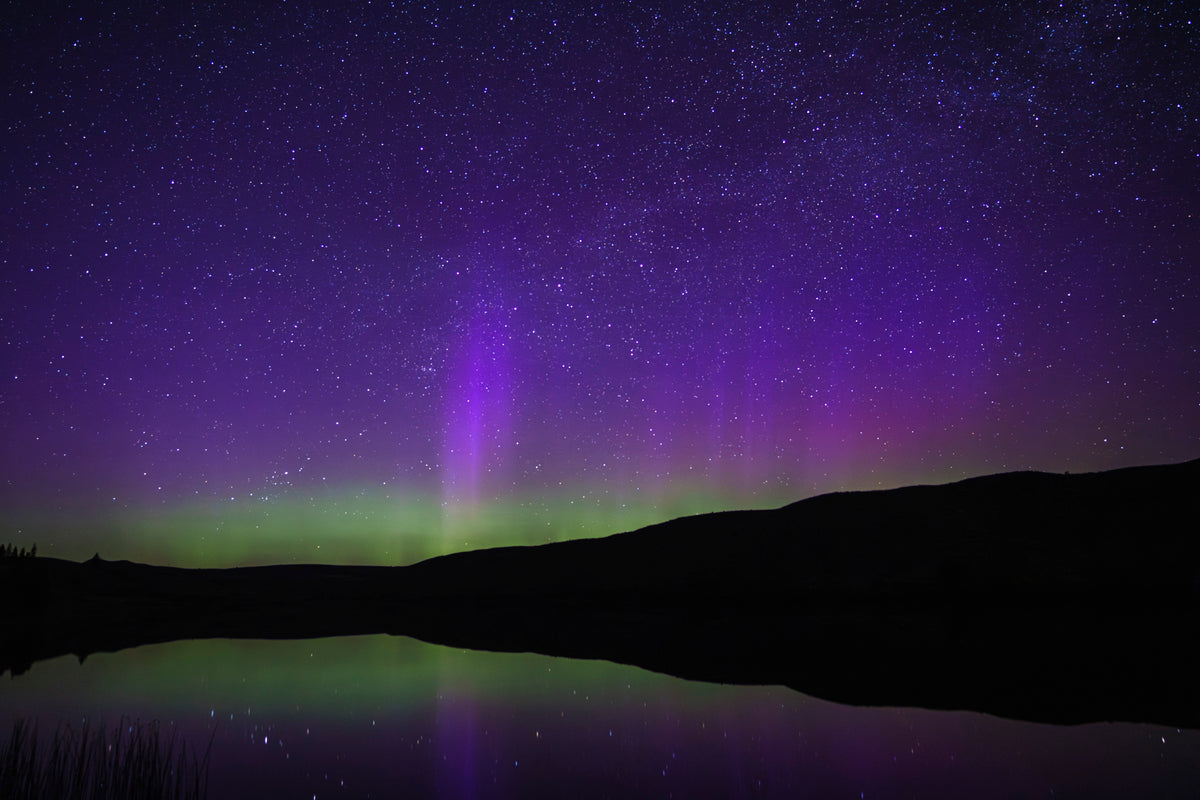 Purple Aurora Reflection - Acrylic Block
2"x3' aspect ratio landscape image shows the aurora borealis reflected on a lake.  The sky above is dark blue and purple, fading to bright green at the horizon.   The horizon is a black line of hills in the foreground, and the lake below the hills is the same purple and green as the sky. There are stars visible in both the sky and the reflection.